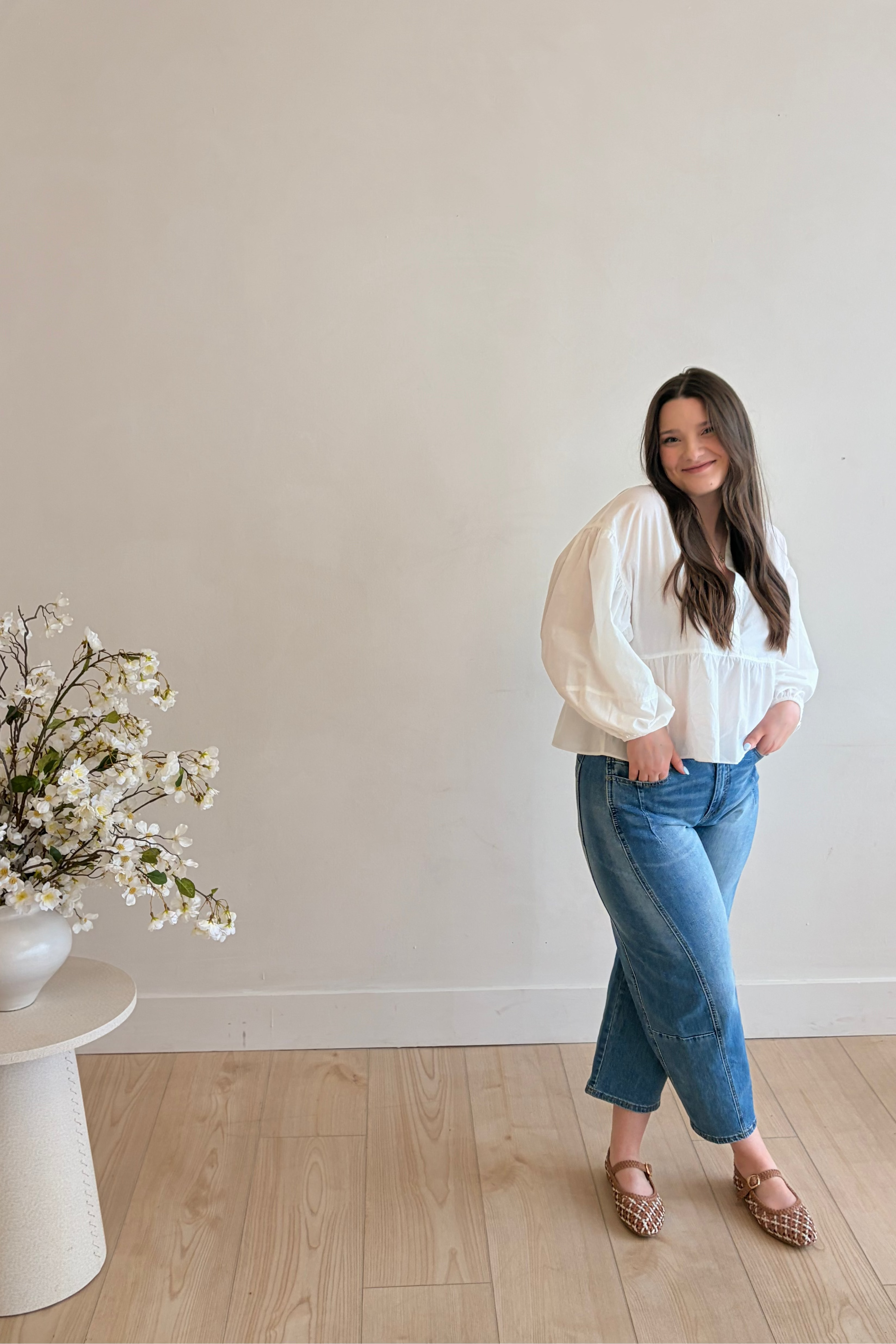 Woman in a white blouse and blue jeans standing in a minimalistic room with a vase of flowers on a small table.