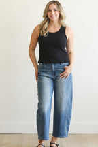 Woman wearing a black tank top and blue jeans against a white wall.