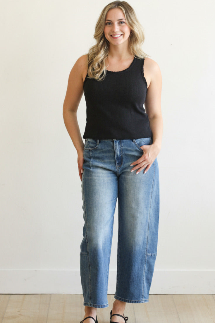 Woman wearing a black tank top and blue jeans against a white wall.